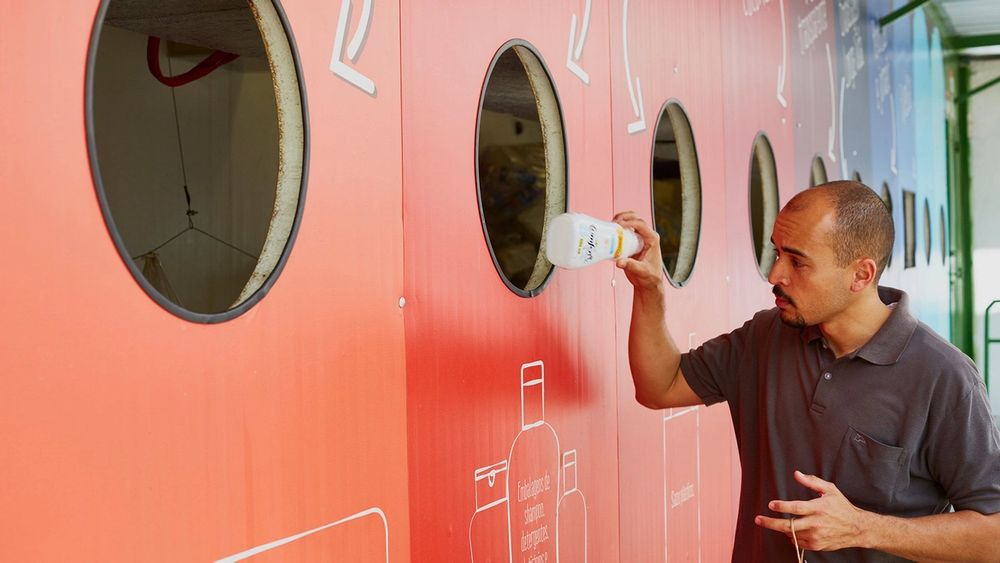 Mann wirft eine Plastikflasche in eine große bunte Recyclingstation.